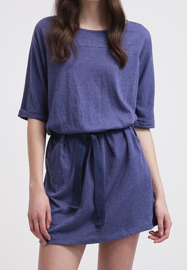 Femme portant une robe bleue décontractée à manches courtes avec une ceinture en tissu nouée et des poches, debout devant un fond clair uni.