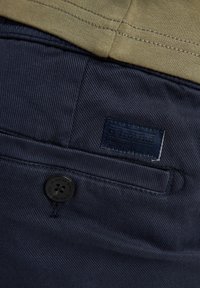 Tissu bleu marine texturé avec des coutures visibles, présentant un bouton noir et une étiquette de la marque Petrol cousue sur un tissu beige au-dessus.