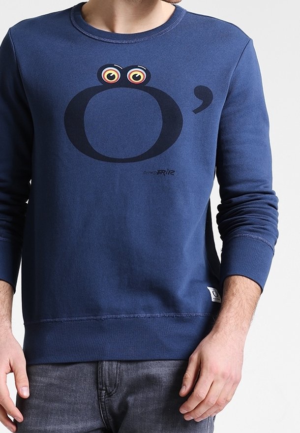 Chemise bleu marine à manches longues avec une grande lettre noire O agrémentée d'yeux de dessin animé au-dessus et d'une petite virgule à droite.
