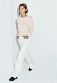 Hellebeige Sweatshirt mit Rundhalsausschnitt und engen Bündchen, kombiniert mit weißen, weiten Jeans; getragen mit schwarzen Schuhen, vor einem neutralen Hintergrund.