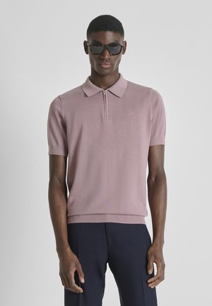 Hombre con gafas de sol negras, camisa polo de manga corta color malva con cremallera y pantalones azul marino oscuro, de pie contra un fondo claro y liso.