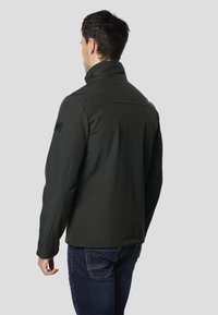 Giacca verde scuro con collo alto, realizzata in un tessuto liscio e strutturato. Presenta cuciture piatte e una toppa con il logo nero sulla spalla sinistra.