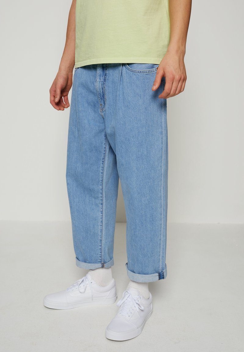 Vaaleansiniset farkut, joissa on väljä istuvuus, käännetyt lahkeet ja klassinen viisi-taskuinen muotoilu, yhdistettynä valkoisiin tennareihin ja lyhyisiin sukkiin.