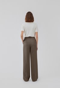 Person with short brown hair stands facing away, wearing a white short-sleeve top tucked into high-waisted wide-leg brown trousers.