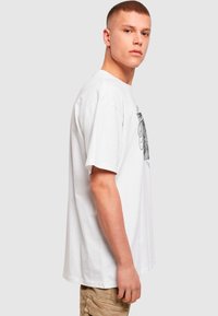 Hvid bomulds t-shirt med et grafisk print på forsiden, korte ærmer og en afslappet pasform. Stoffet ser blødt og glat ud.