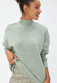 Jasnozielony sweter typu golf wykonan z miękkiego dzianinowego materiału, z prążkowaną teksturą przy szyi i mankietach, w połączeniu z kraciastą spódnicą.