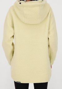 Fleecejacka i ljusgul färg med texturerad yta, huva och något förlängd rygg. Märkesetikett synlig nära fållen.