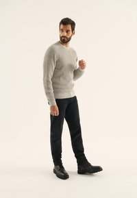 Suéter gris con canalé y mangas largas, pantalones negros ajustados y botas negras hasta el tobillo. La modelo está de pie con una expresión neutra contra un fondo claro.