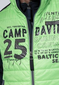 Zielona pikowana kurtka puchowa z wyraźnym czarnym nadrukiem tekstu i cyfr, zamkiem błyskawicznym oraz kontrastowymi ciemnymi akcentami na ramionach.