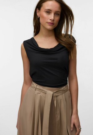 Kvinde med langt brunt hår iført en ærmeløs sort top og en beige nederdel med høj talje og bindebælte, poserer mod en hvid baggrund.