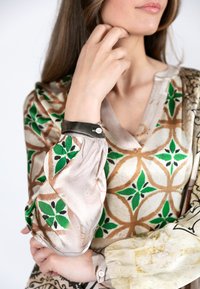 Seidenbluse mit geometrischem Muster in grünen und braunen Farbtönen. Die Ärmel sind mit einem dunklen Akzentbund und Knopfdetail gerafft.
