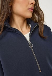Felpa blu navy con zip, caratterizzata da un colletto alto, una zip metallica con un tirante rotondo e un tessuto morbido e texturizzato. Inquadratura ravvicinata del collo.
