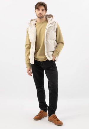 Mann trägt beige gesteppte Weste, beigefarbenen Sweatshirt, schwarze Jeans und braune Turnschuhe und steht vor weißem Hintergrund.