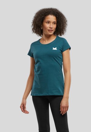Kobieta z kręconymi włosami, ubrana w turkusowy t-shirt z krótkim rękawem z małym białym motywem kota na piersi oraz czarne legginsy, stoi na szarym tle.