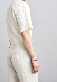 Mies, joka on pukeutunut teksturoituun vaaleankerman väriseen lyhythihasa paita ja vaaleanbeigeihin housuihin, näkyy takaa ja sivulta, ranteessa on mustasta helmistä tehty rannekoru.