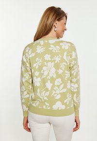 Strickpullover in hellgrün mit weißem Blumenmuster, gerippten Bündchen und Saum sowie Rundhalsausschnitt. Das Modell trägt ihn mit weißen Hosen.
