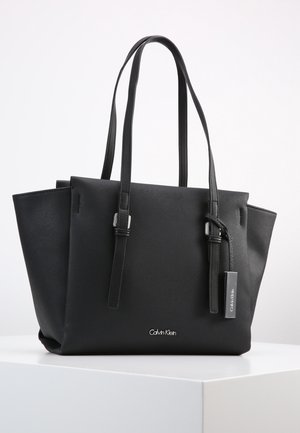 Schwarze Calvin Klein Tote-Bag mit doppelten Schulterriemen, strukturierten Seiten und einem metallenen Marken-Logo-Anhänger, der an einem Riemen hängt.