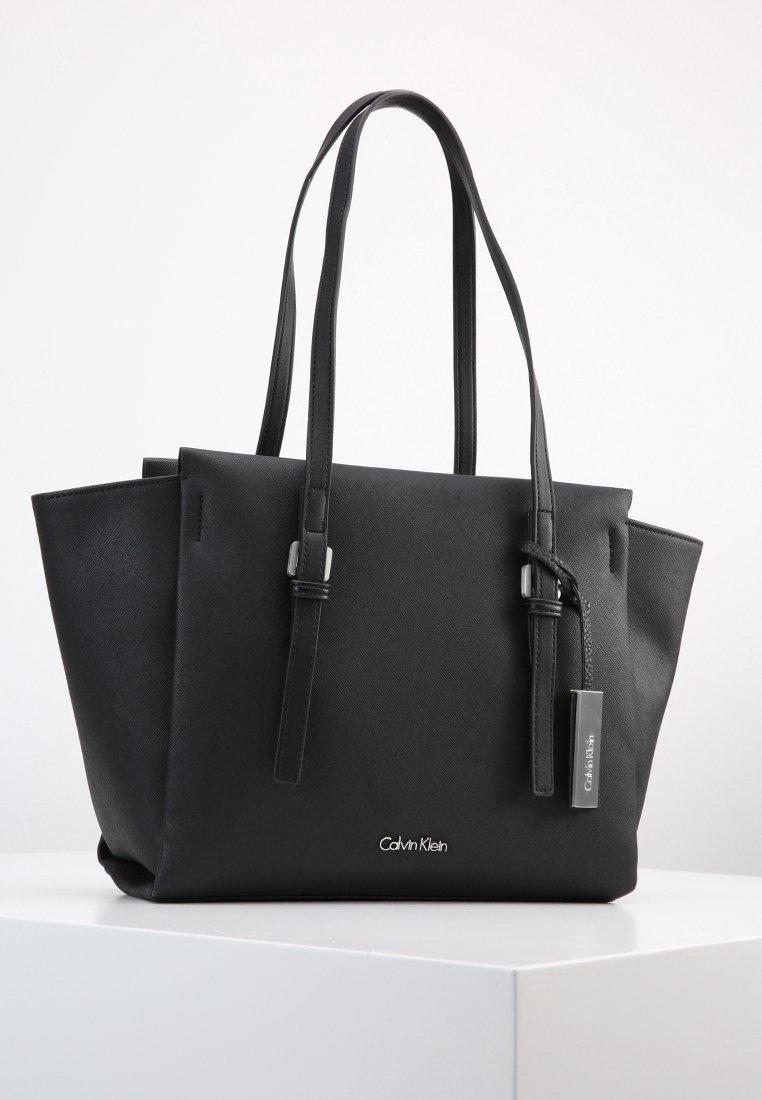 Sac cabas noir Calvin Klein avec double anse d'épaule, côtés structurés et une étiquette métallique à logo de marque suspendue à une anse.