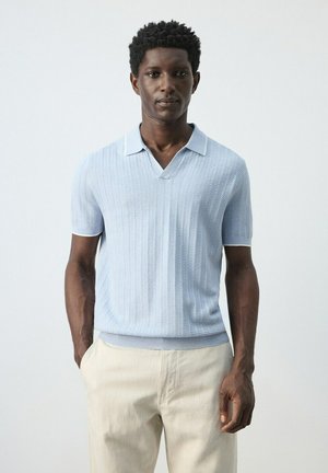 Hombre con camisa polo de manga corta texturizada azul claro y pantalones beige, de pie frente a un fondo blanco liso.