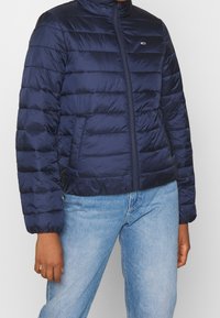 Personne portant une veste matelassée bleu marine fermée, associée à un jean en denim bleu clair, se tenant avec les bras détendus sur les côtés.