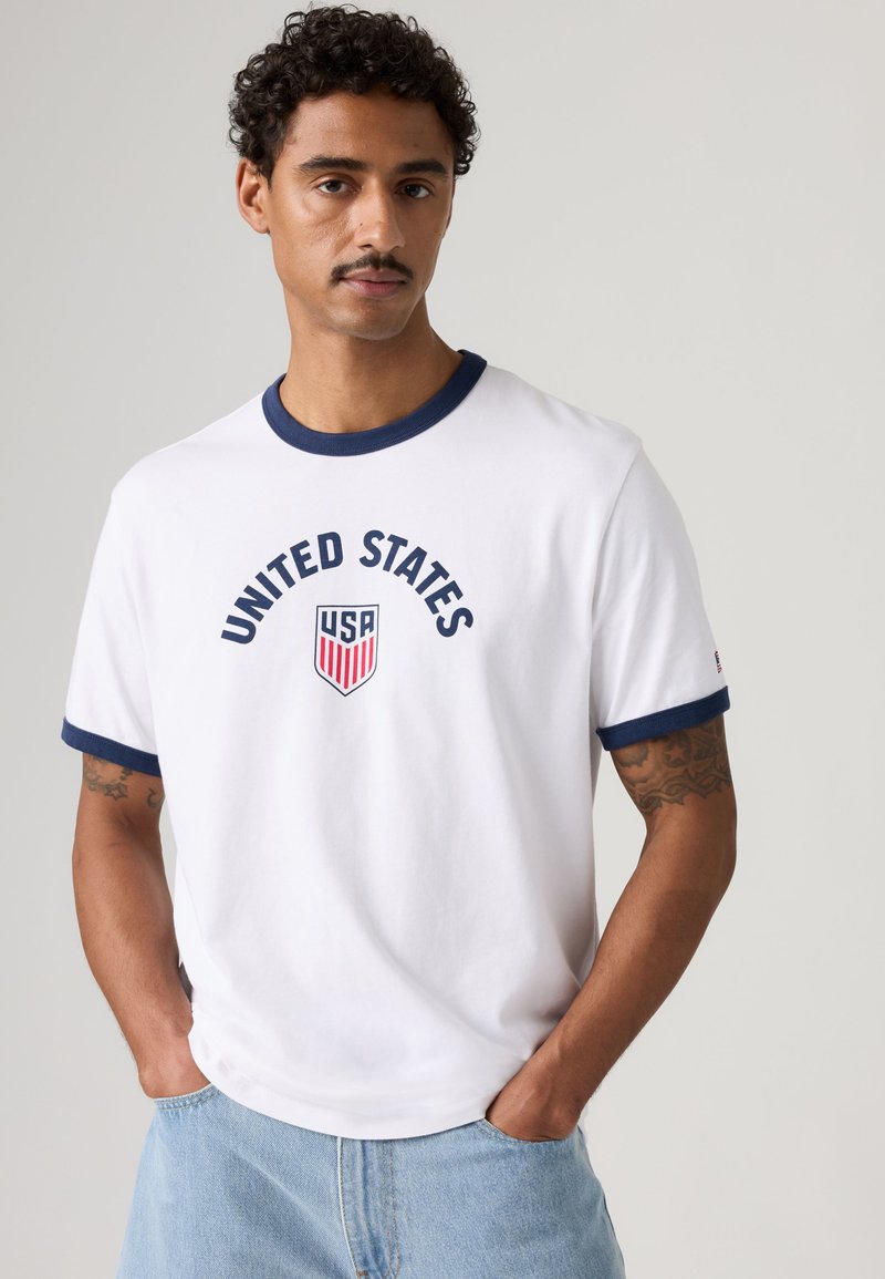 Man with curly hair and mustache wears white T-shirt with navy trim and "UNITED STATES" and USA shield logo, hands in light blue jeans pockets.