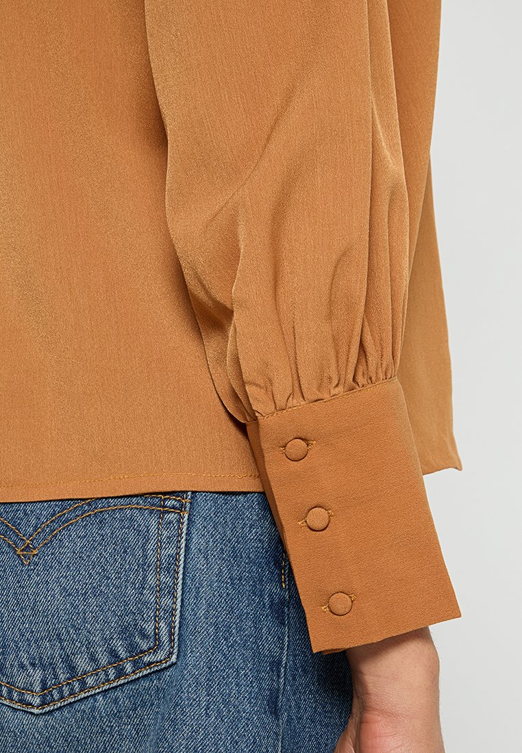 Mouw van een bruine blouse met drie met stof bedekte knopen op de manchetten, gecombineerd met blauwe denim jeans met stiksel op de achterzak.