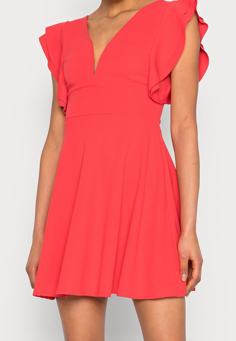 Robe rouge sans manches avec un décolleté en V profond et des détails volantés aux épaules, portée par une personne debout montrée du cou jusqu'à mi-cuisse.