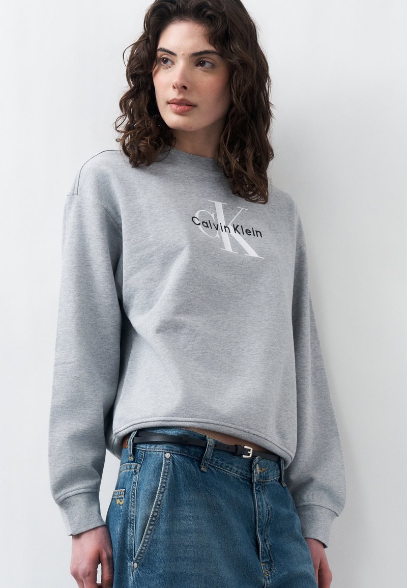 Jeune femme aux cheveux bouclés portant un sweat-shirt gris Calvin Klein et un jean bleu, debout devant un fond blanc uni.