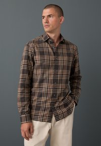 Camicia a quadri marrone e beige realizzata in tessuto con colletto abbottonato, maniche lunghe e orlo arrotondato; caratteristiche bottoni scuri.