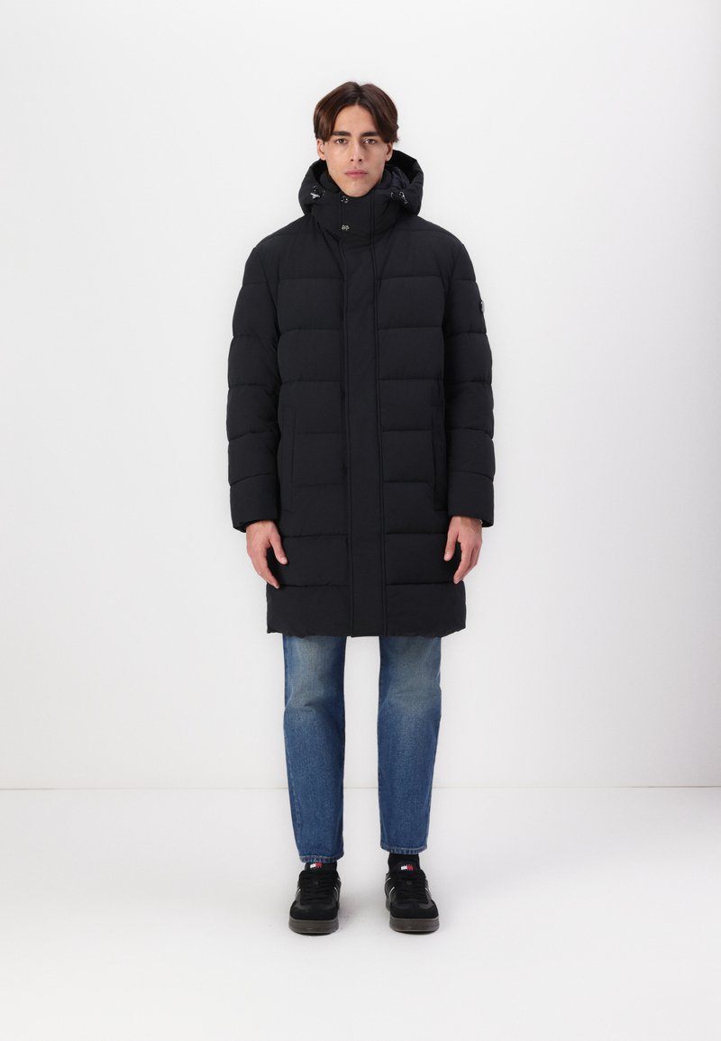 Manteau rembourré noir avec un col haut et une capuche, doté d'une fermeture éclair à l'avant et d'un bouton, porté avec un jean en denim bleu et des chaussures noires.