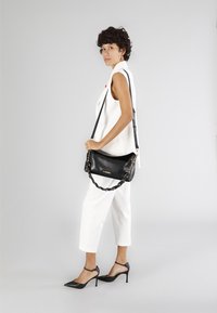 Sort læderhåndtaske med kæde og stofrem, glat tekstur og guld logo. Båret crossbody med et hvidt outfit og sorte hæle.