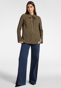 Donna che indossa un blouse a maniche lunghe verde oliva con dettagli a fiocco e borchie, abbinato a jeans a gamba larga blu scuro e scarpe marroni.
