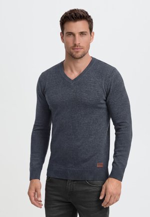 Homme portant un pull gris foncé ajusté à col en V et un pantalon noir, debout devant un fond clair uni, regardant devant lui.