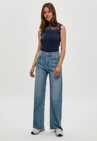 Navy Tanktop mit Spitzenbesatz, kombiniert mit hellblauen weit geschnittenen Jeans und grauen Sneakers. Glatte Stoffqualität, lockere Passform und fransiger Saum.