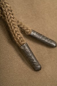 Cuerda trenzada de color marrón con terminales metálicos plateados grabados con la palabra "Reell", sobre un fondo de tela beige texturizada, destacando los detalles de la artesanía.