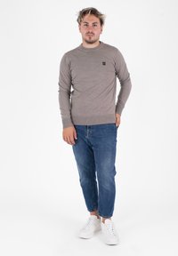 Maglione lavorato a maglia color marrone chiaro con scollo rotondo e maniche lunghe, abbinato a jeans blu e sneaker bianche. Piccolo logo sul maglione.
