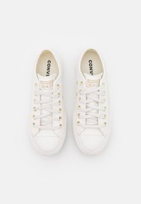Fehér vászon sneaker arany fűzőlyukakkal és fűzőkkel, kerek orral, diszkrét márkalogóval a nyelvén és texturált gumitalppal.