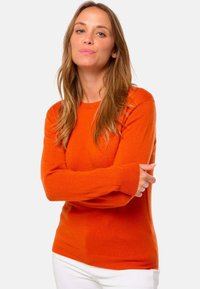 Pull en tricot orange vif, avec un col rond et des manches longues à poignets côtelés. Porté avec un pantalon blanc.