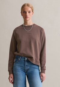 Felpa marrone con una vestibilità leggermente rilassata e maniche raglan, abbinata a jeans blu a vita alta. Presenta un logo minimal al centro.