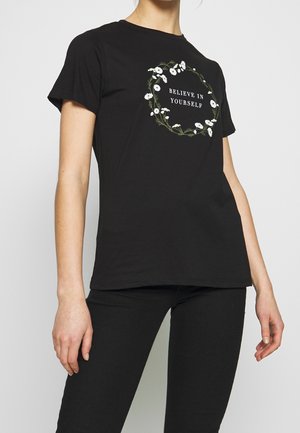 Osoba nosząca czarną koszulkę z białym wieńcem stokrotek otaczającym napis „WIERZ W SIEBIE” na piersi, w połączeniu z czarnymi spodniami.