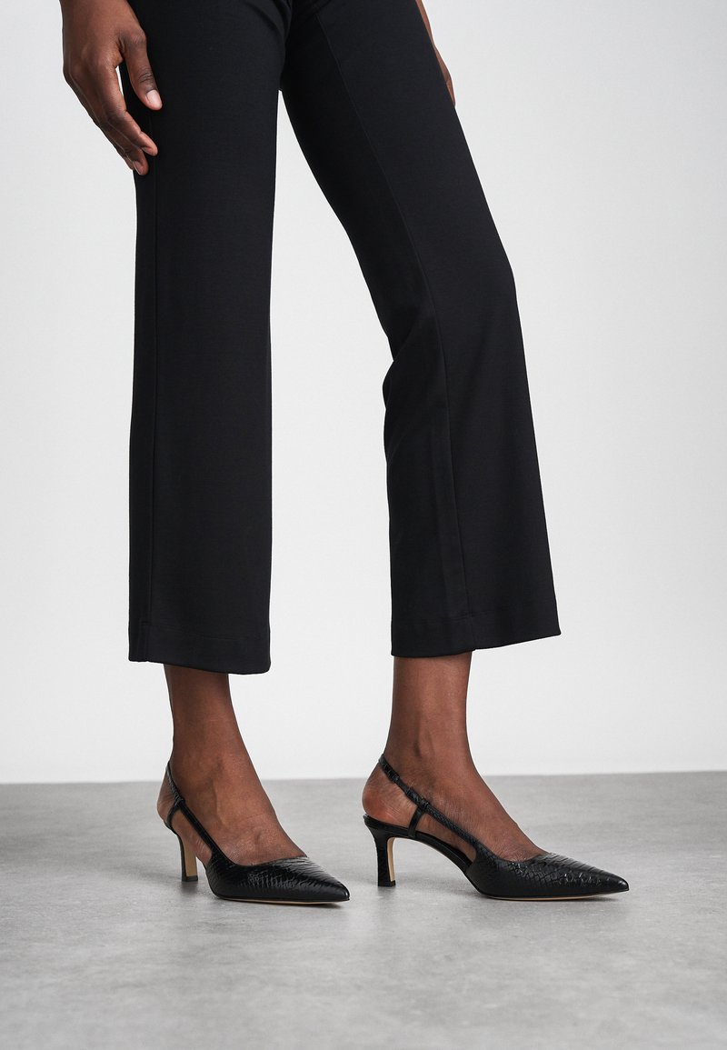 Schwarze Slingback-Absätze mit spitzem Zehenbereich, strukturiertem Reptilmuster und dünnem Absatz. Getragen mit cropped schwarzen Hosen auf einer neutralen Oberfläche.