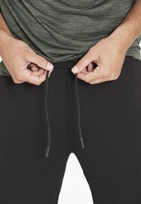 Svarta joggerbyxor med dragsko i midjan, som visar en texturerad grön skjorta ovanför. Dragskon har svarta ändar; tyget verkar mjukt och flexibelt.