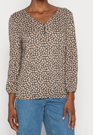 Blouse beige à manches longues avec un motif géométrique marron foncé, décolleté en V avec patte de boutonnage et poignets élastiques.