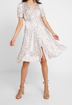 Kobieta w białej kwiatowej sukience z dekoltem w serek i bufkami na rękawach, nosząca beżowe sandały na szerokim obcasie z paskami, sukienka lekko podniesiona, ukazująca rozcięcie na nodze.