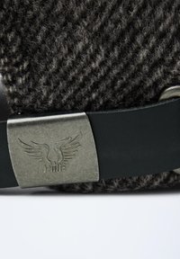 Gros plan sur une boucle en métal avec un logo ailé fixée à un tissu texturé noir et une sangle noire.