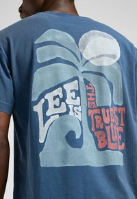 Sininen puuvillapaita, jossa on graafinen kuvasto palmusta, aalloista ja teksti "Lee on totisin sininen" kermansävyisenä ja punaisena. Pehmeä rakenne.