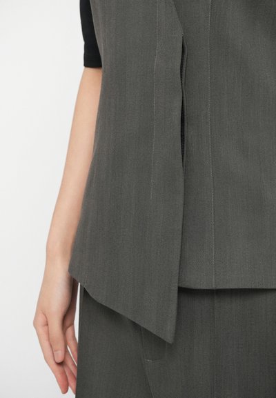 Sivý жилет s texturovanou látkou, so priamej strihom a šikmým spodným okrajom, navrstvený na čiernom topu. Ruka umiestnená na boku.