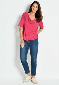 Ein pinkfarbener, drappierter Top mit kurzen Ärmeln, kombiniert mit blauen Denim-Jeans. Das Model trägt cremefarbene Sneakers. Weicher Stoff mit lässigem Schnitt.