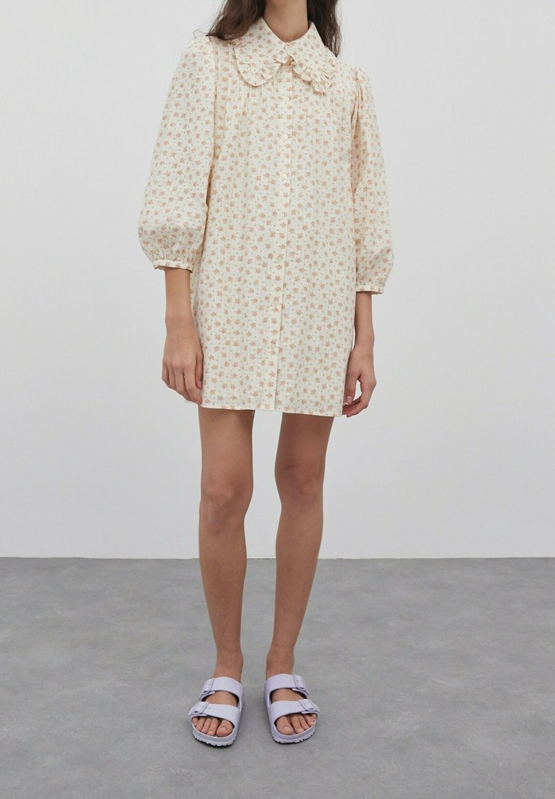Femme portant une robe crème à motifs floraux avec des manches bouffantes et un grand col, associée à des sandales lilas à boucle sur un sol gris.