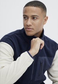 Veste en polaire avec des blocs de couleurs bleu marine et crème. Col haut, poche poitrine zippée, tissu texturé avec des accents lisses et bords pipés.